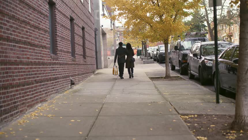 A man and a woman walk down a street passing by trees with brown leaves and a row of cars