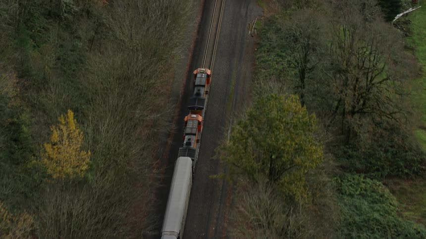 Aerial following two orange and black engines pulling a train along a tree lined track through the countryside beside a wooded area a little before nightfall on a fall day