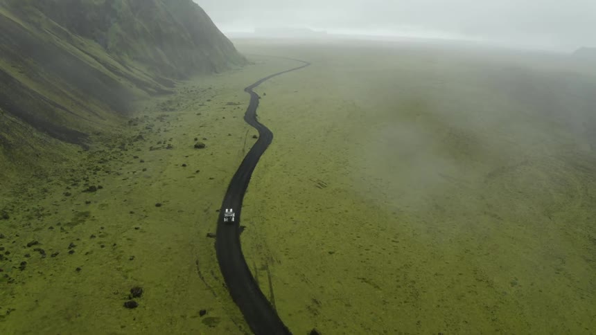 White suv driving along a dark black road surrounded by bright green grass on a misty day