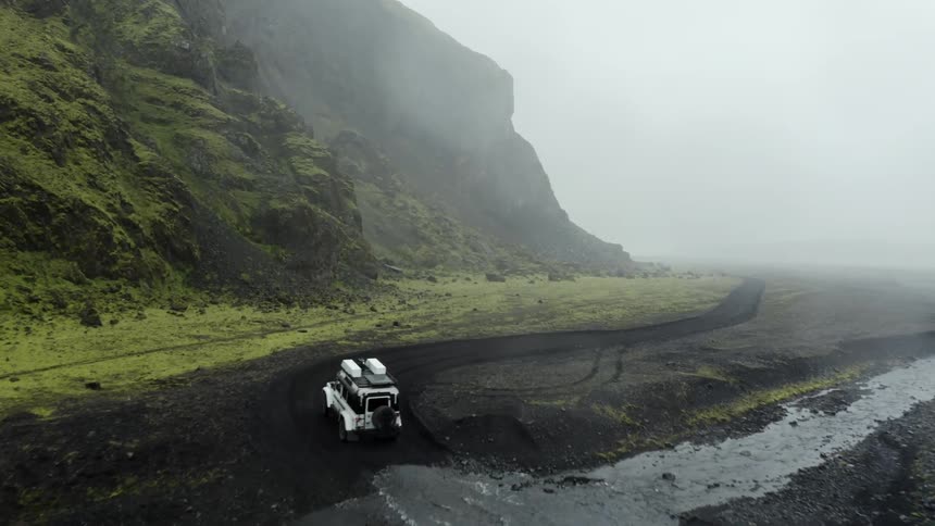 Jeep driving through puddles on a dirt road next to mountains with coniferous trees on a hazy day