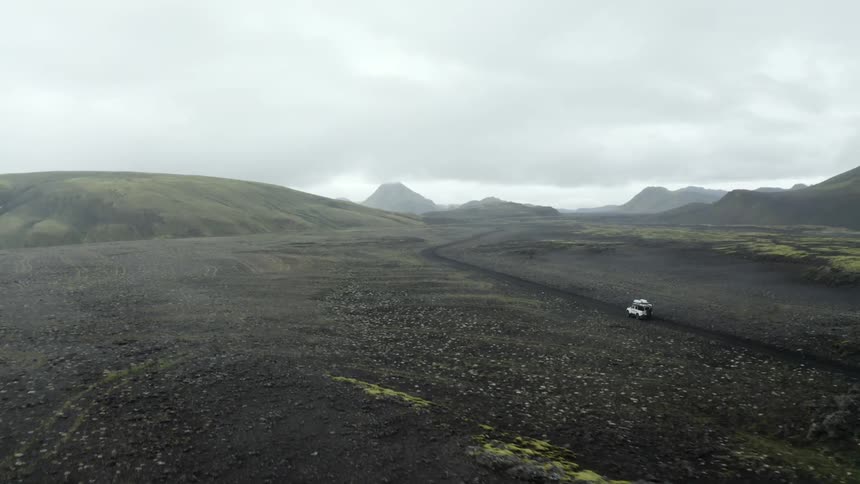 Vehicle drives along dirt road through plain bordering hills on an overcast day