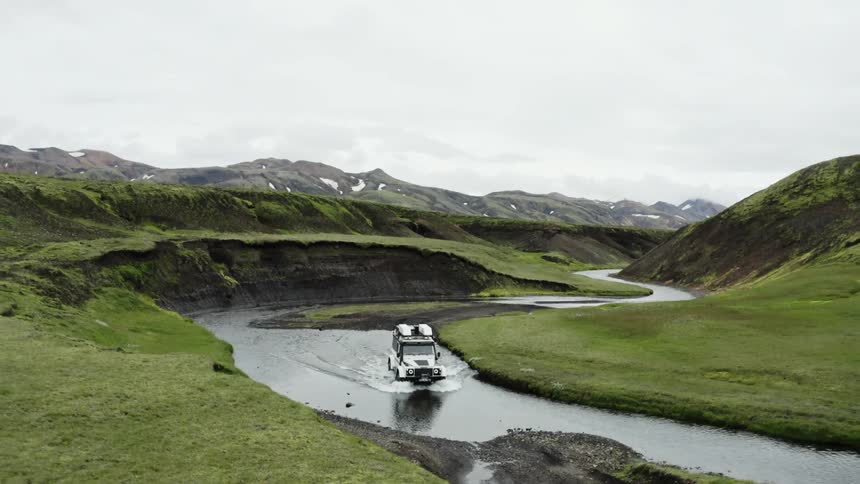 Vehicle fords stream separating segments of road through hilly terrain bordered by mountains on an overcast day