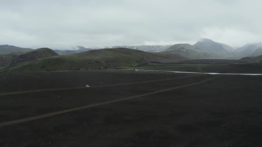 Vehicle drives along road intersecting stream with mountains in the background