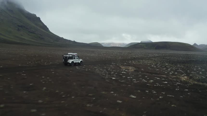 An offroad vehicle traverses a desolate rocky landscape with mountains in the background under a cloudy sky