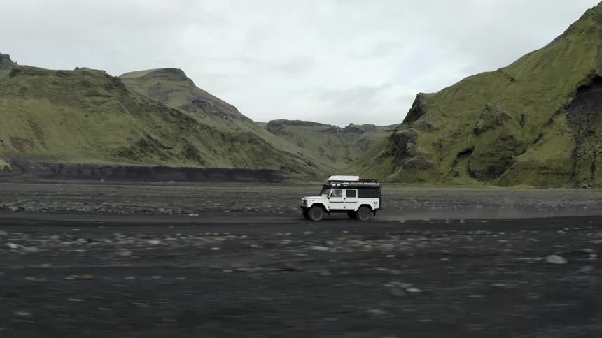 A white suv car driving on a ground with black sand and grassy hills in the background