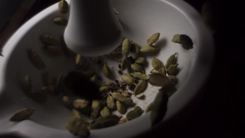 A white ceramic pestle crushing cardamom seeds in a mortar in slow motion