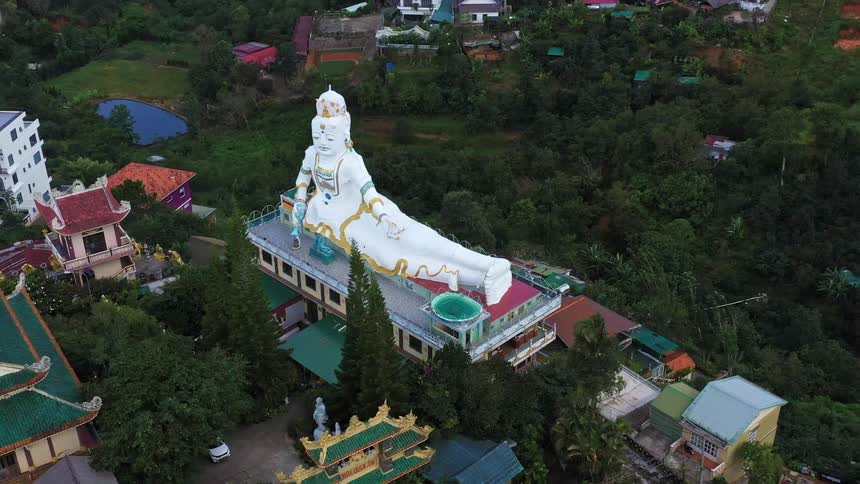 Aerial of giant white buddha laying with legs extended on to the side on top of a pagoda with other smaller pagodas and lush green trees around it in vietnam