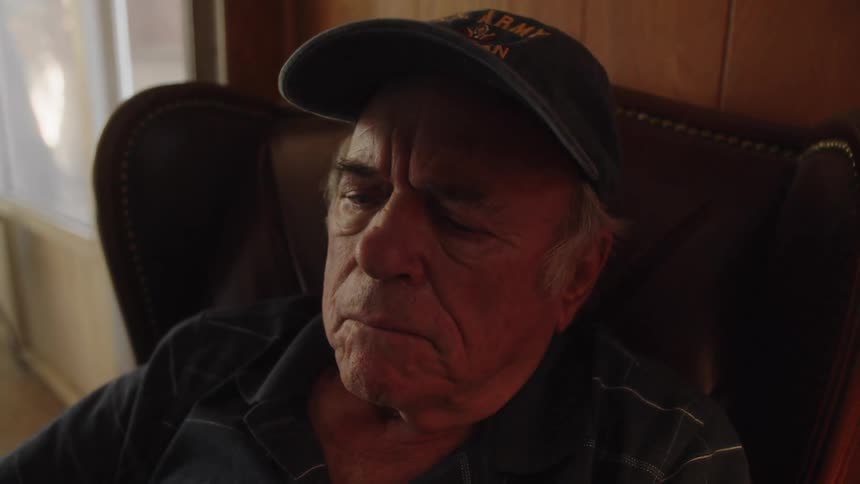 Elderly man wearing a cap seated in an armchair putting on his glasses