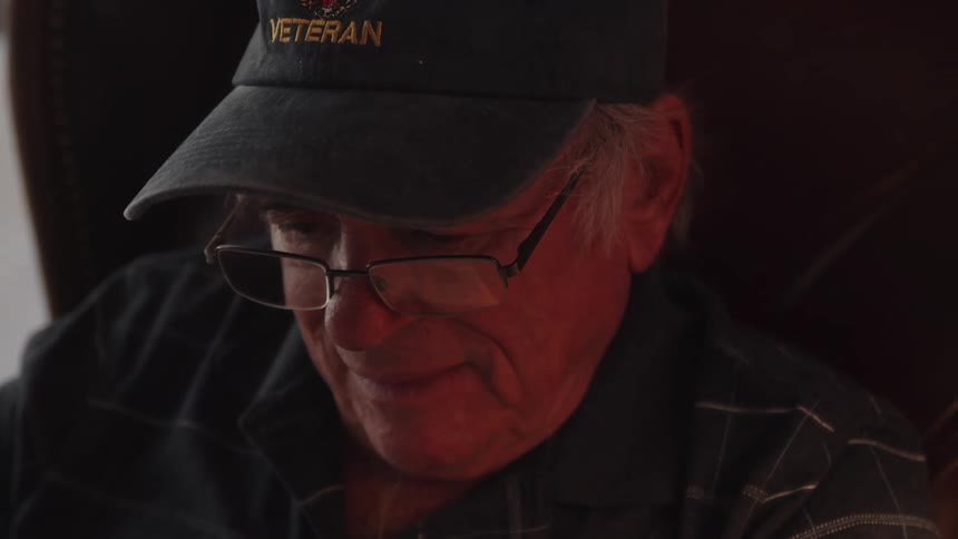 An elderly man wearing a veteran cap a dark blue shirt and glasses sits in an armchair reading