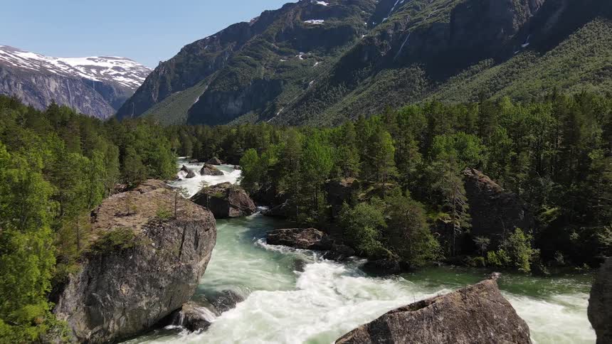 A rugged river flowing through a forested valley with large boulders and mountainous backdrop