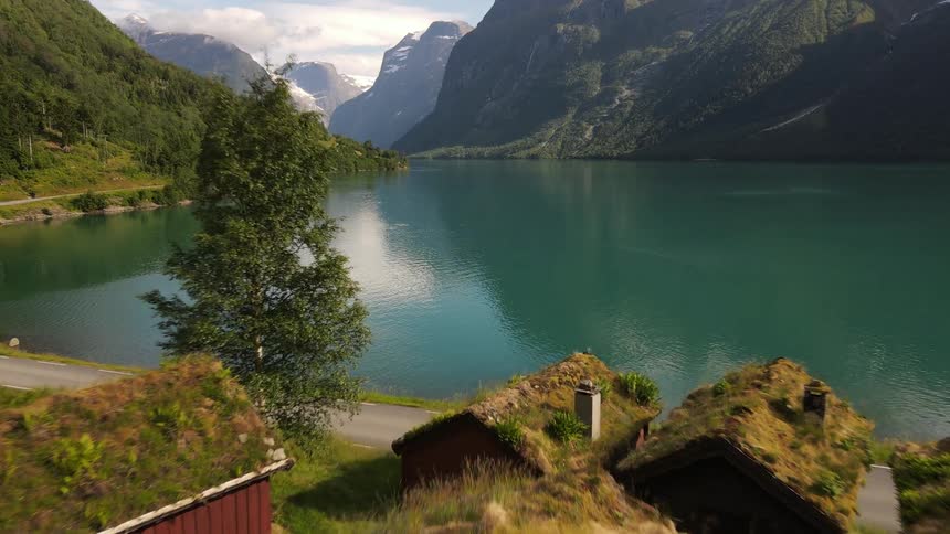 Decent down the green hillside past cabins with moss roofs to a lake that reflects the clouds and blue sky and is surrounded by mountains