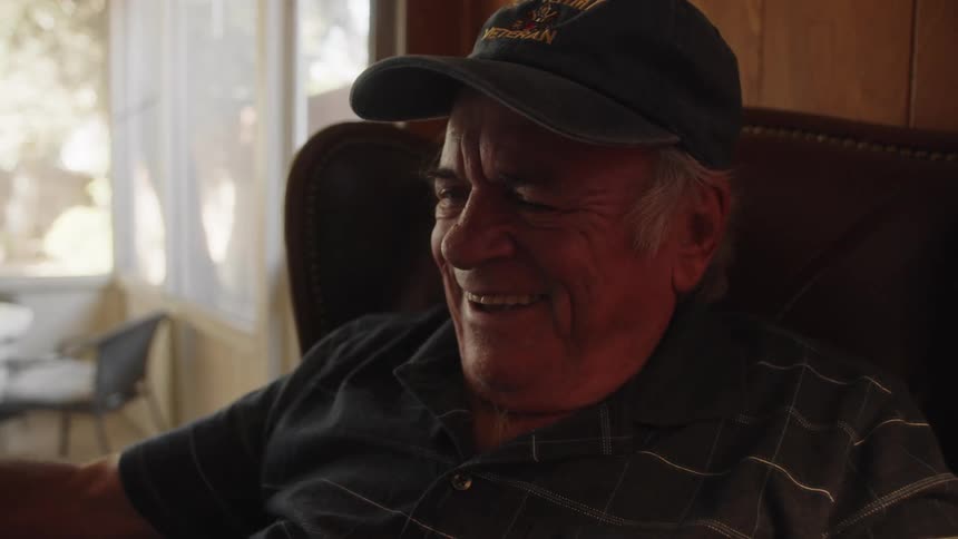 Elderly man wearing a veteran cap and blue shirt sits in armchair laughing out