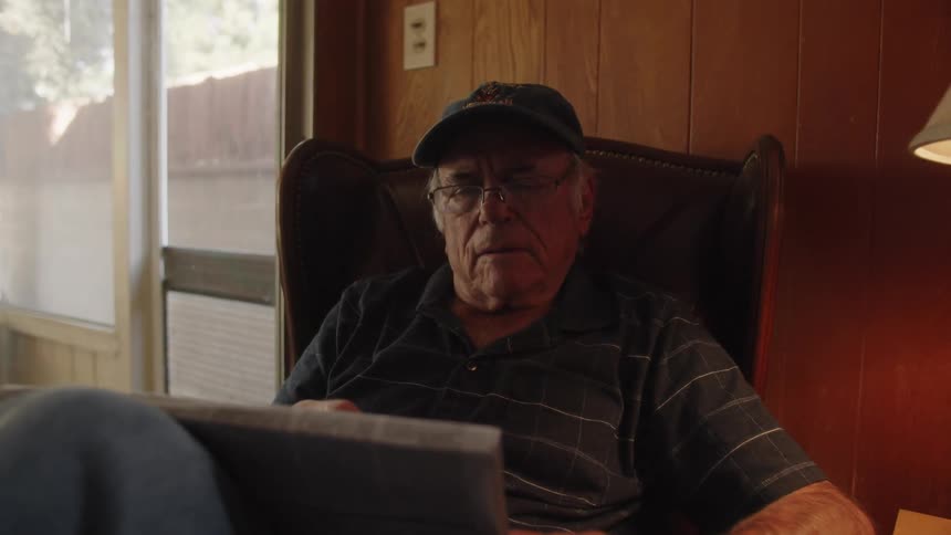 An elderly man reading a newspaper while seated on a chair indoor
