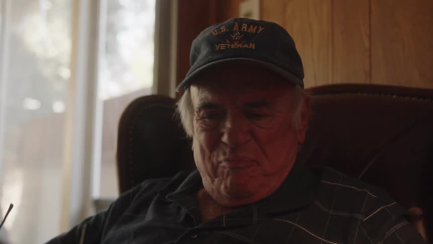 An elderly man is sitting in a chair reading as he laughs and takes off his glasses and reaches for a can and drinks