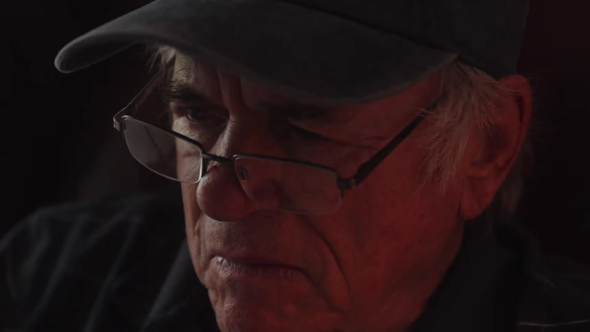 An elderly man wearing a baseball cap and glasses concentrates