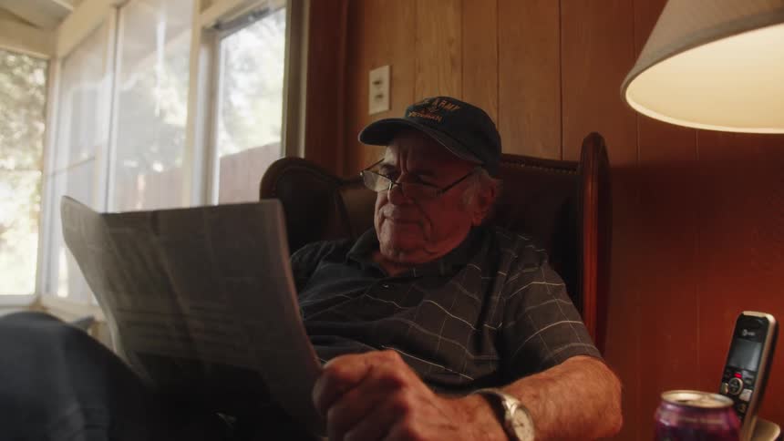 Elderly man wearing a cap and a t shirt reads newspaper in room