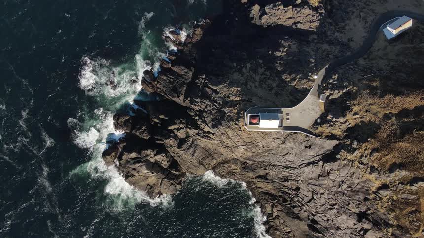 Aerial view from above of a lighthouse on a rocky promontory and the ocean waves splashing into the shoreline rocks