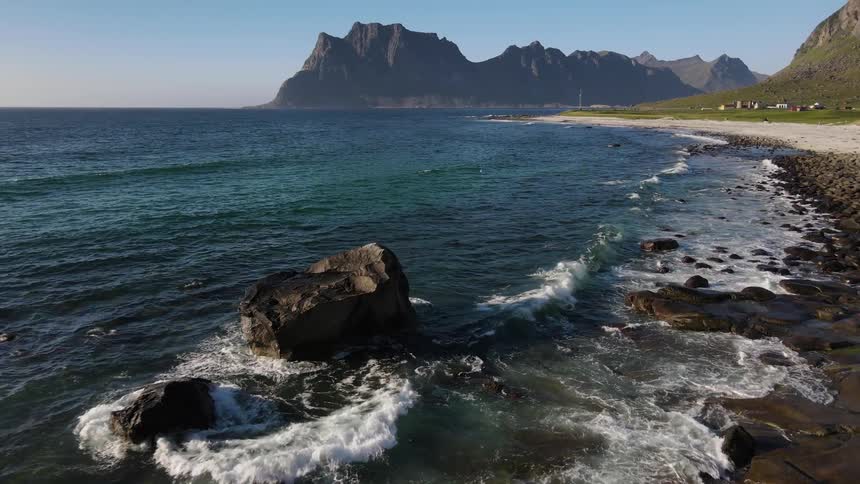 Waves roll into shore and splash against rocks along the coast with mountains in the distance