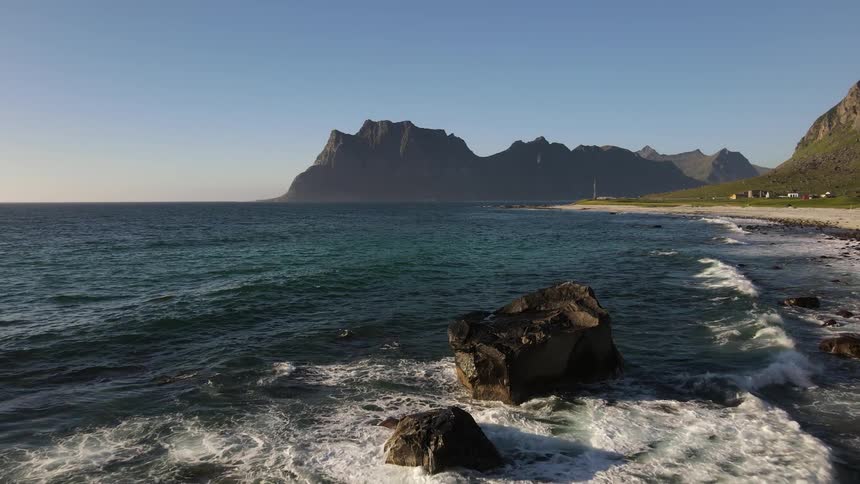 Waves roll into shore and splash against rocks along the coast with mountains in the distance
