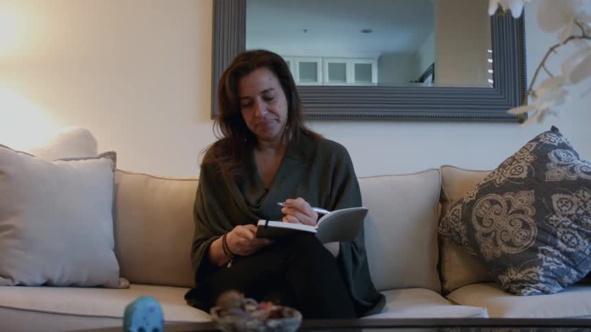 A woman walks over and sits on a couch writing in a book