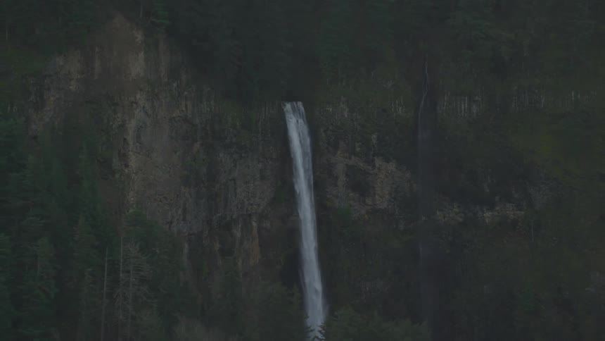 Aerial of a waterfall on the side of a woodland tree covered mountain after sundown