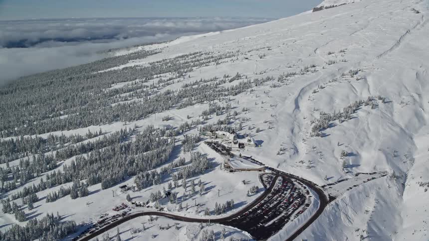 An aerial view of buildings and a parking lot located on a snowy mountain slope near a forest