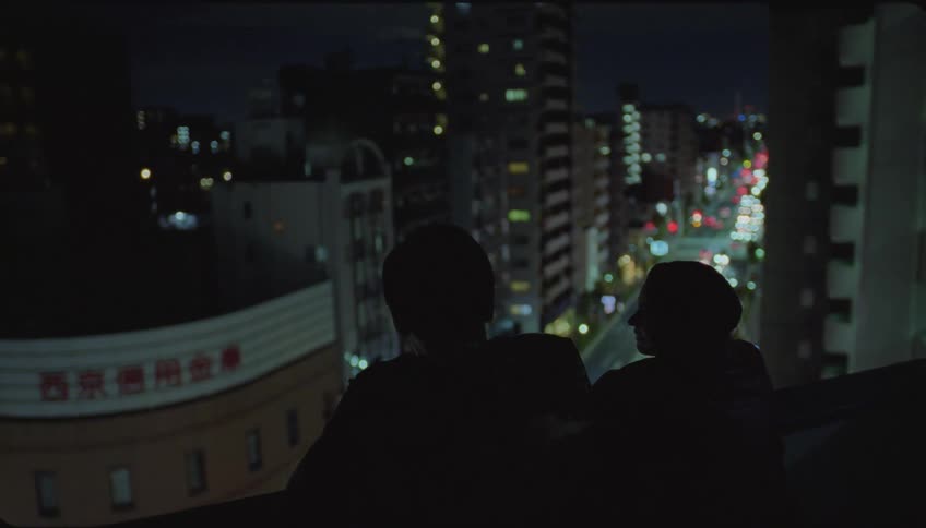 Two people talking while standing at the top of a city building at night