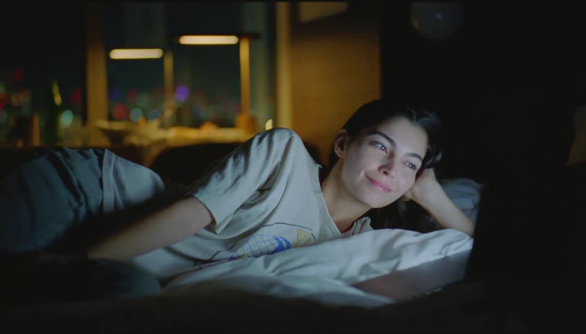 A woman is watching something on her laptop in the dark and laughs