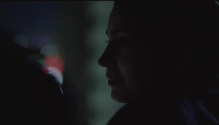 A woman with earrings and dark hair talks to another woman watching the city skyline through a window at night