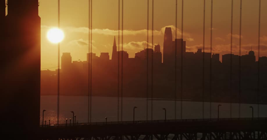 View through a suspension bridge of the sun glowing on the horizon behind the metropolitan city skyline