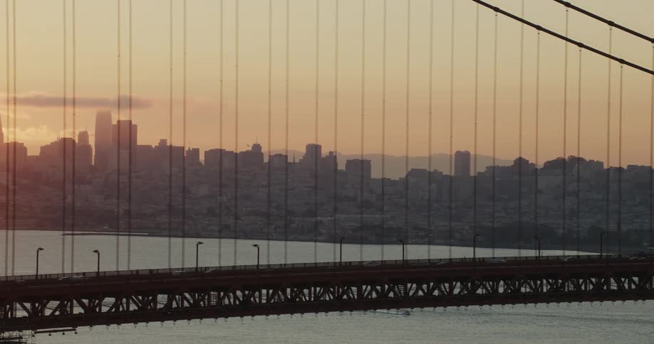 Suspension bridge section over the bay with the city skyline covered in an orange haze and the sun rising in the sun in the background