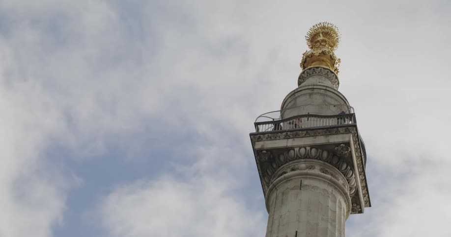 Monument to the great fire in london top portion of pedestal with gold statue top as clouds move overhead on a sunny day