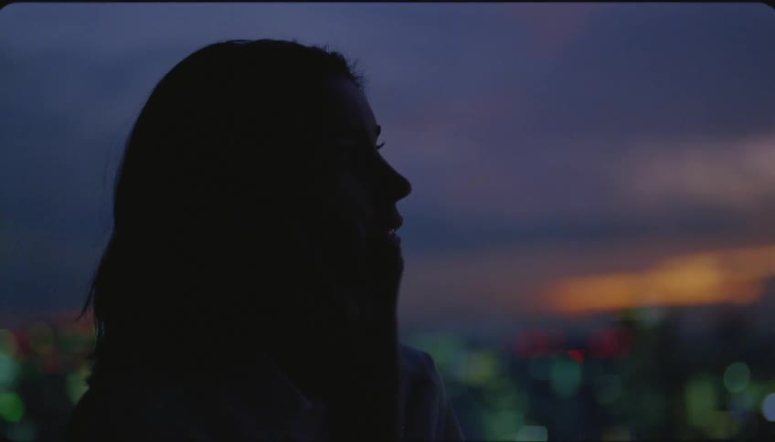 Woman talking on smartphone is silhouetted against the city skyline and sky at dusk
