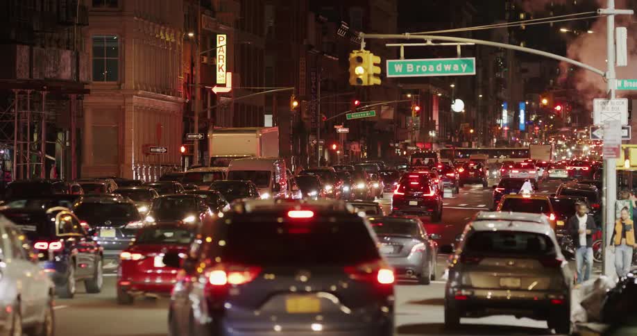 A bustling new york city street illuminated at night with numerous cars in traffic and pedestrians on the sidewalk