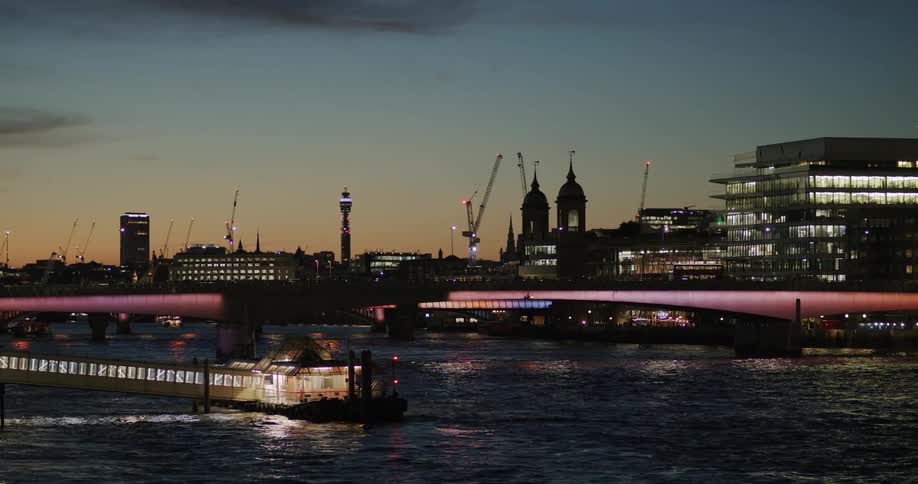 A well lit riverboat and a bridge across a river with a silhouetted buildings against a dull orange and blue sky at sunset