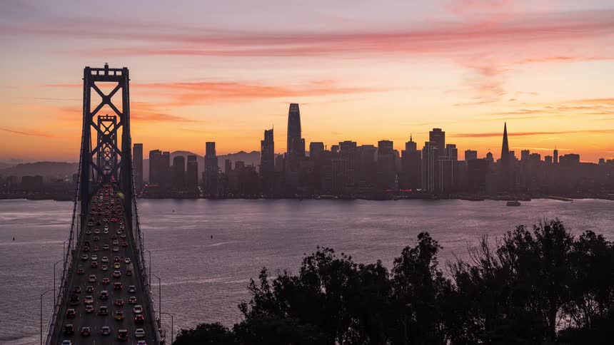 Timelapse of San Francisco an the sun setting on the horizon with a landscape of a bridge over the sea with vehicles heading towards a metropolis