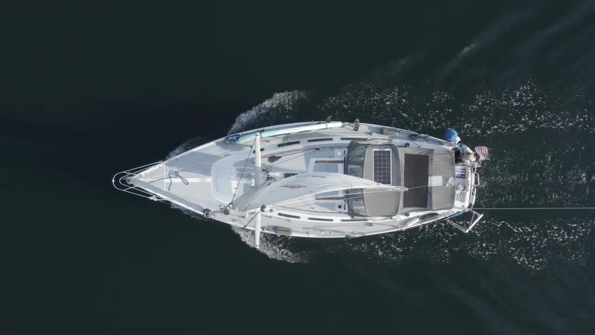 Aerial over a white boat with a white white sail moving through dark water during the day