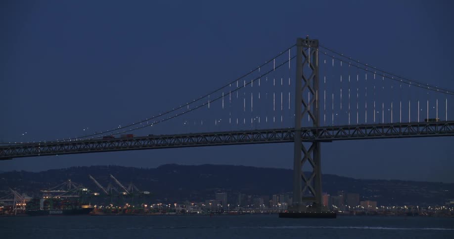 Suspension bridge over the bay and the shoreline lit up with cranes in front of mountain range and dark blue sky at night