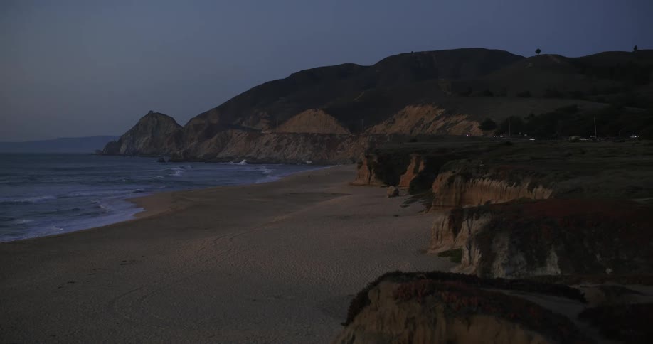 Ocean waves roll into the beach with cars travelling on a road in the distance