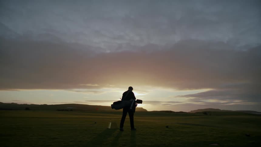 Golfer carrying his golf bag walking across the green grass of a golf course silhouetted against the bright horizon at sunset