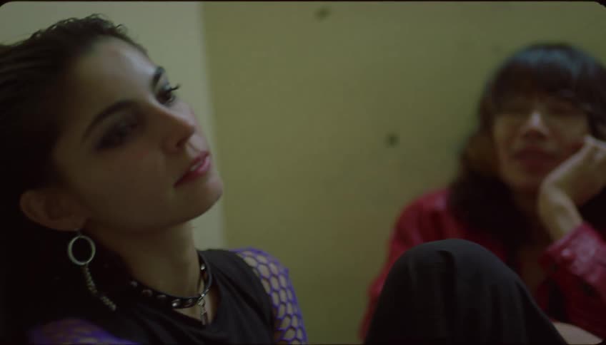 Two people are waiting at a yellow wall the man yawns and rests his head on the womans shoulder the camera pans to show another woman who laughs