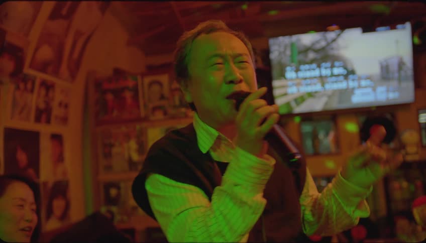 A man is happily singing karaoke and some people are sitting there clapping for him
