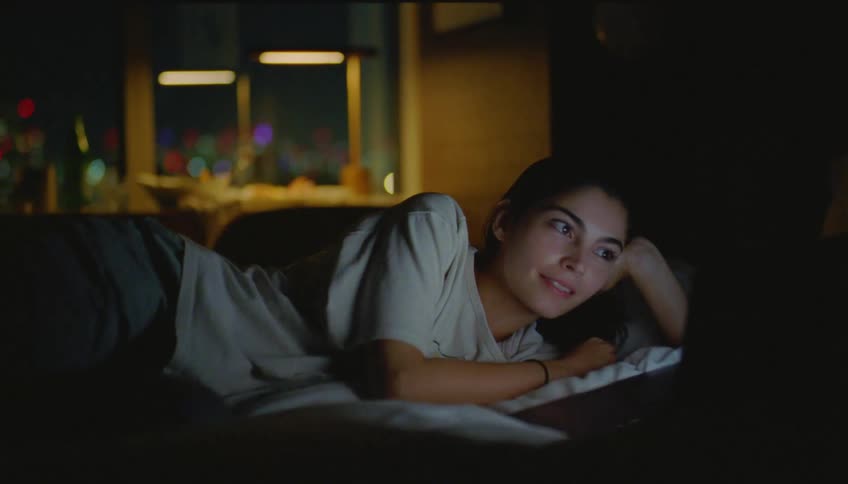 Woman wearing a white t shirt and pants watch movie on laptop lying on bed at night