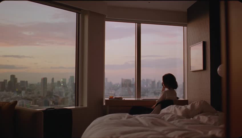 A woman sits at the desk in front of the bedroom window talking on the phone while gazing at the dawn view of the city