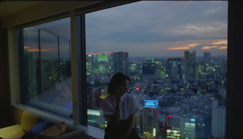 Woman sits near window overlooking city at night and talks on smartphone