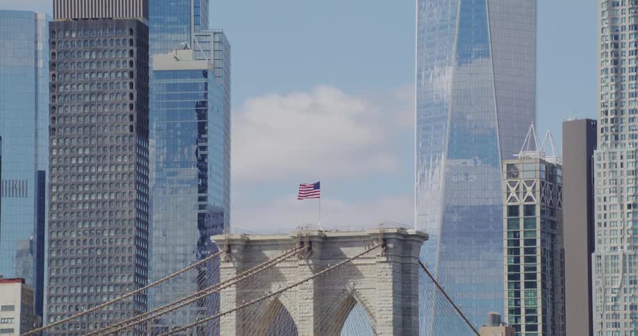 One world trade center towers over skyscrapers near american flag topped bridge on a cloudy day