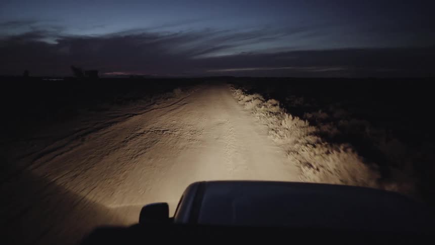 A vehicle's headlights illuminate a rural dirt road as it drives at night