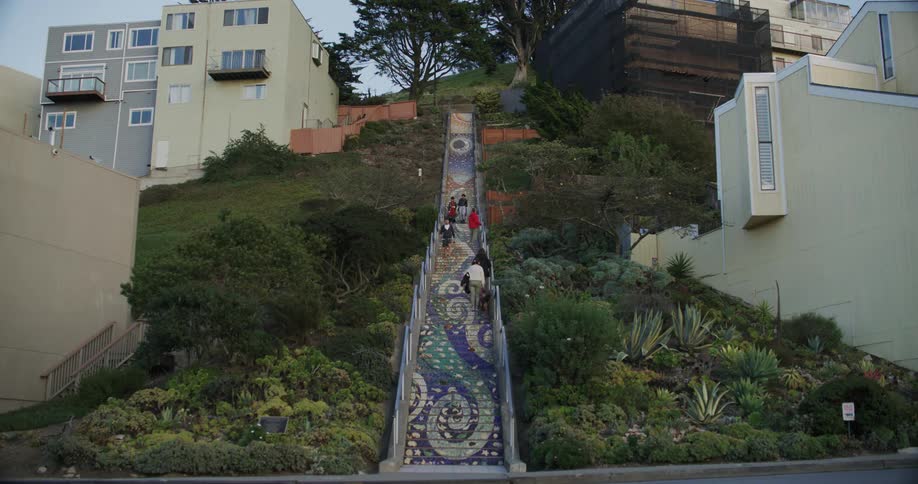 People are climbing a tall run of outdoor stairs with abstract designs up the side of a hill with condo buildings at the top as cars pass by on the road below