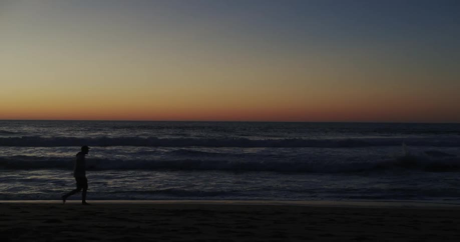 A man chases his dog on the beach as waves roll in under a sunset sky