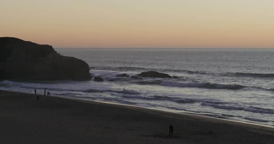People walk on the beach with their pet dog as waves roll into shore and splash against rocks along the coast